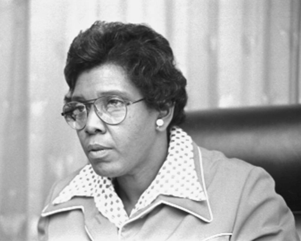 A head-and-shoulders photograph of Congresswoman Barbara Jordan, a Black woman. Jordan is seated in a Congressional chamber in a leather chair facing the left side of the screen, and is wearing glasses and a polka dot collared blouse with a jacket over it. Image source: Library of Congress